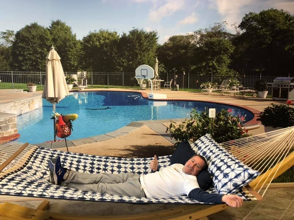 Man relaxes in hammock by pool; blue water, trees, and patio in the background.