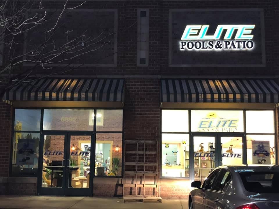 Elite Pools & Patio storefront at night, lit signage above entrance, awning over windows. A parked car is in front.
