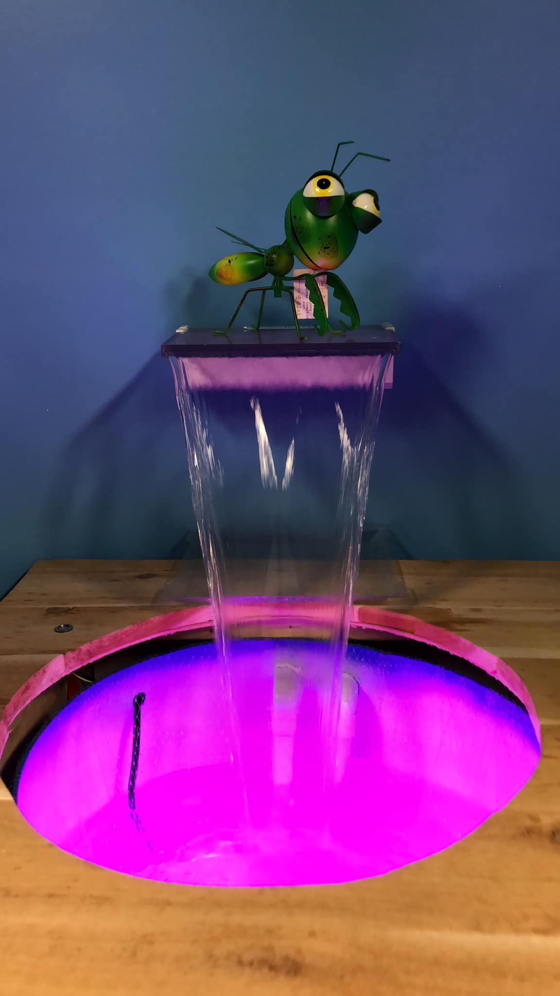 Green ant statue on a waterfall fountain with purple lights, set on a wood surface, against a blue wall.