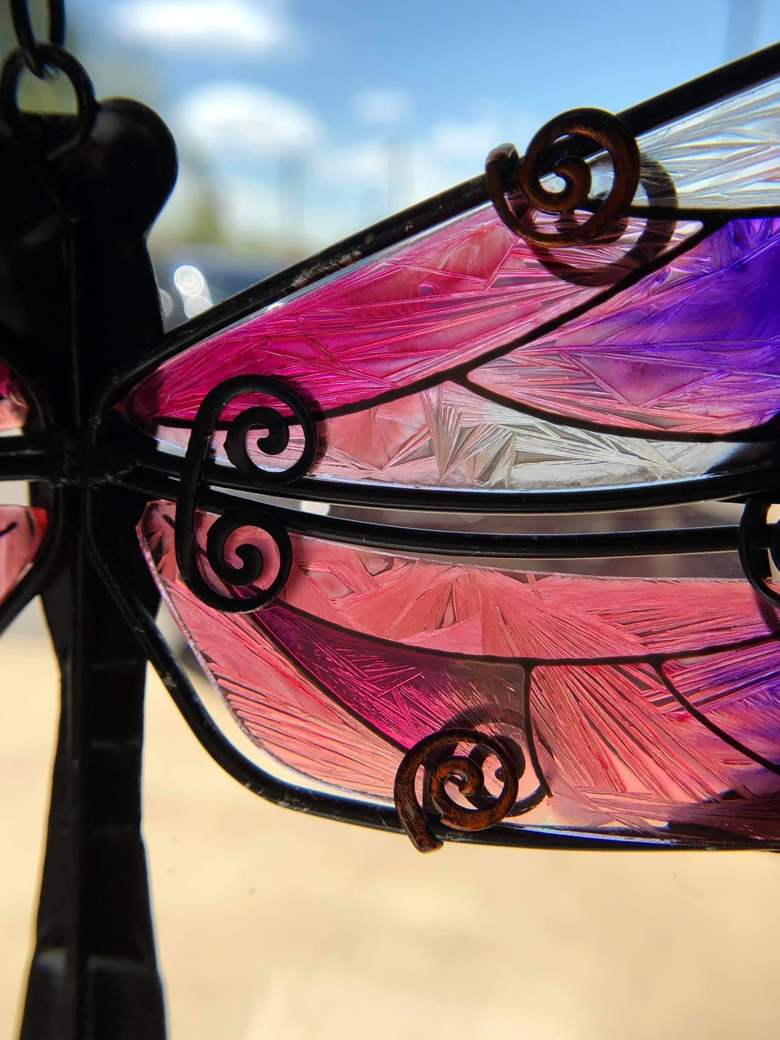 Stained glass dragonfly with pink and purple wings, metallic details, and a black frame.