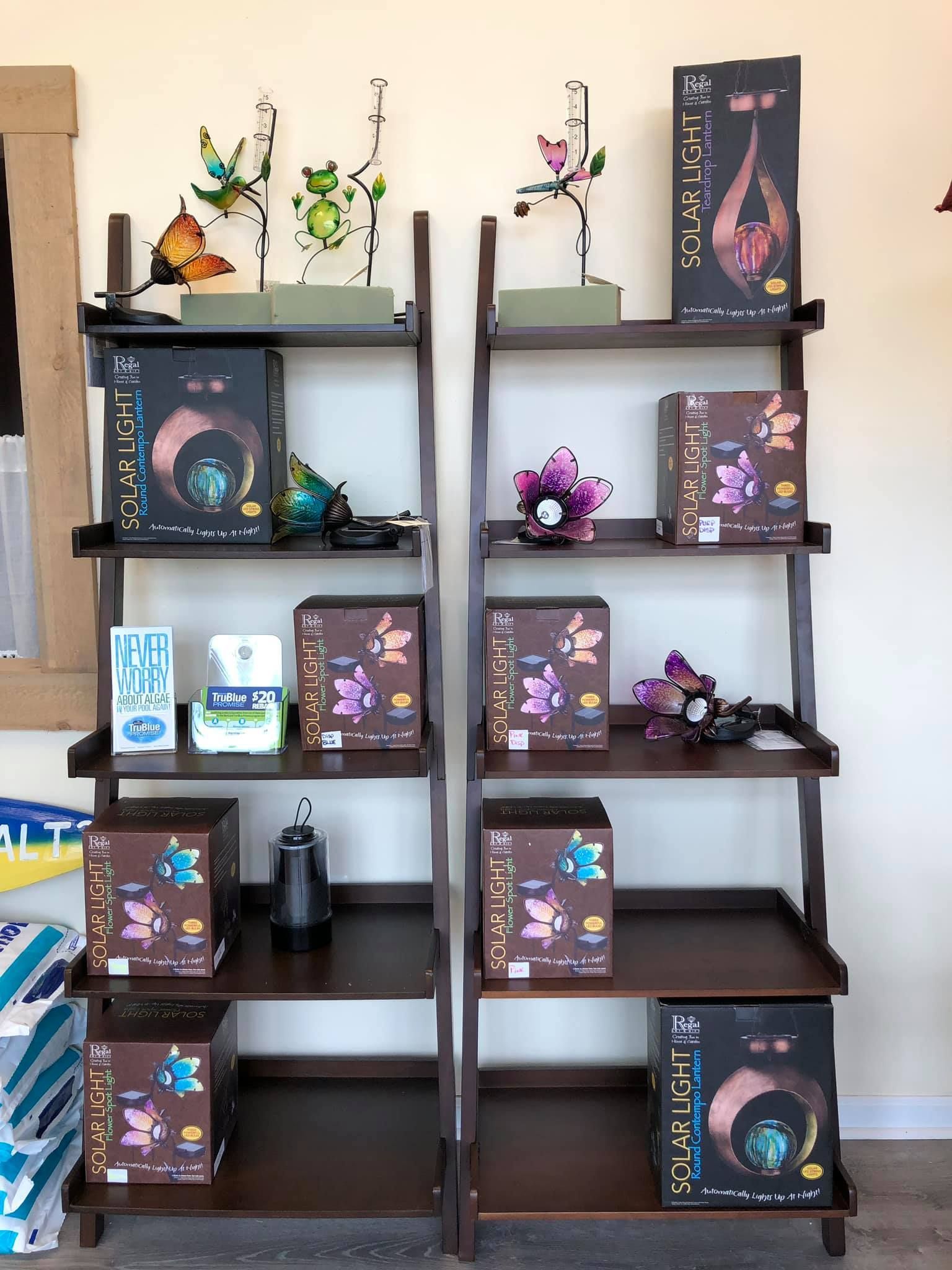 Two wooden leaning shelves displaying boxes, decorative plants, and a small dark-colored appliance.
