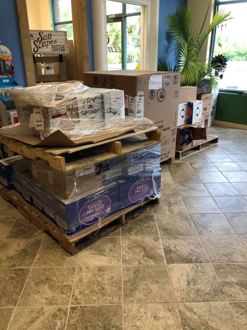 Pallets of wrapped boxes inside a store. Brown tiled floor, tall plant, and blue and white walls visible.