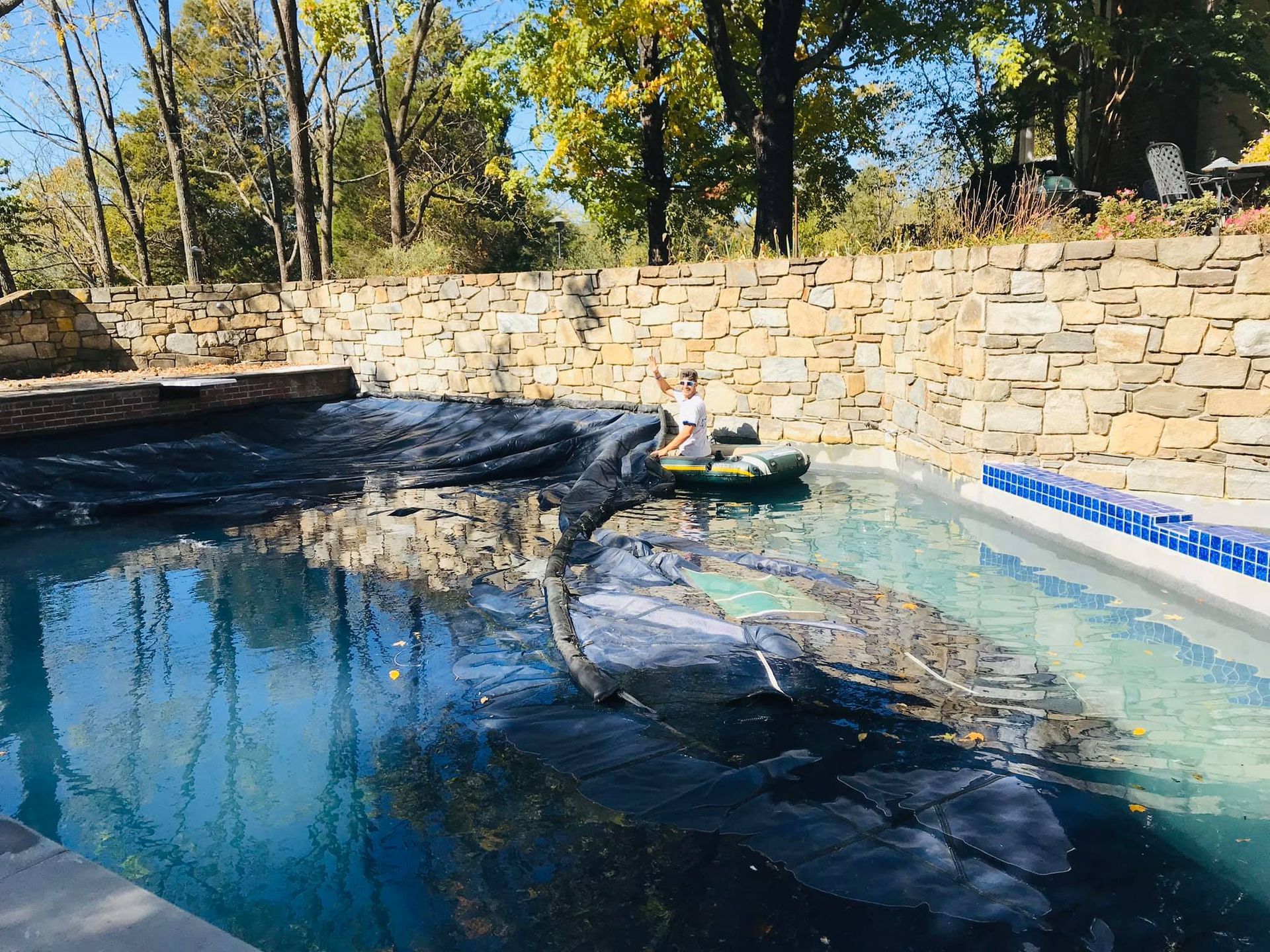 Person in small boat cleaning debris from a pool with a stone retaining wall and trees in the background.