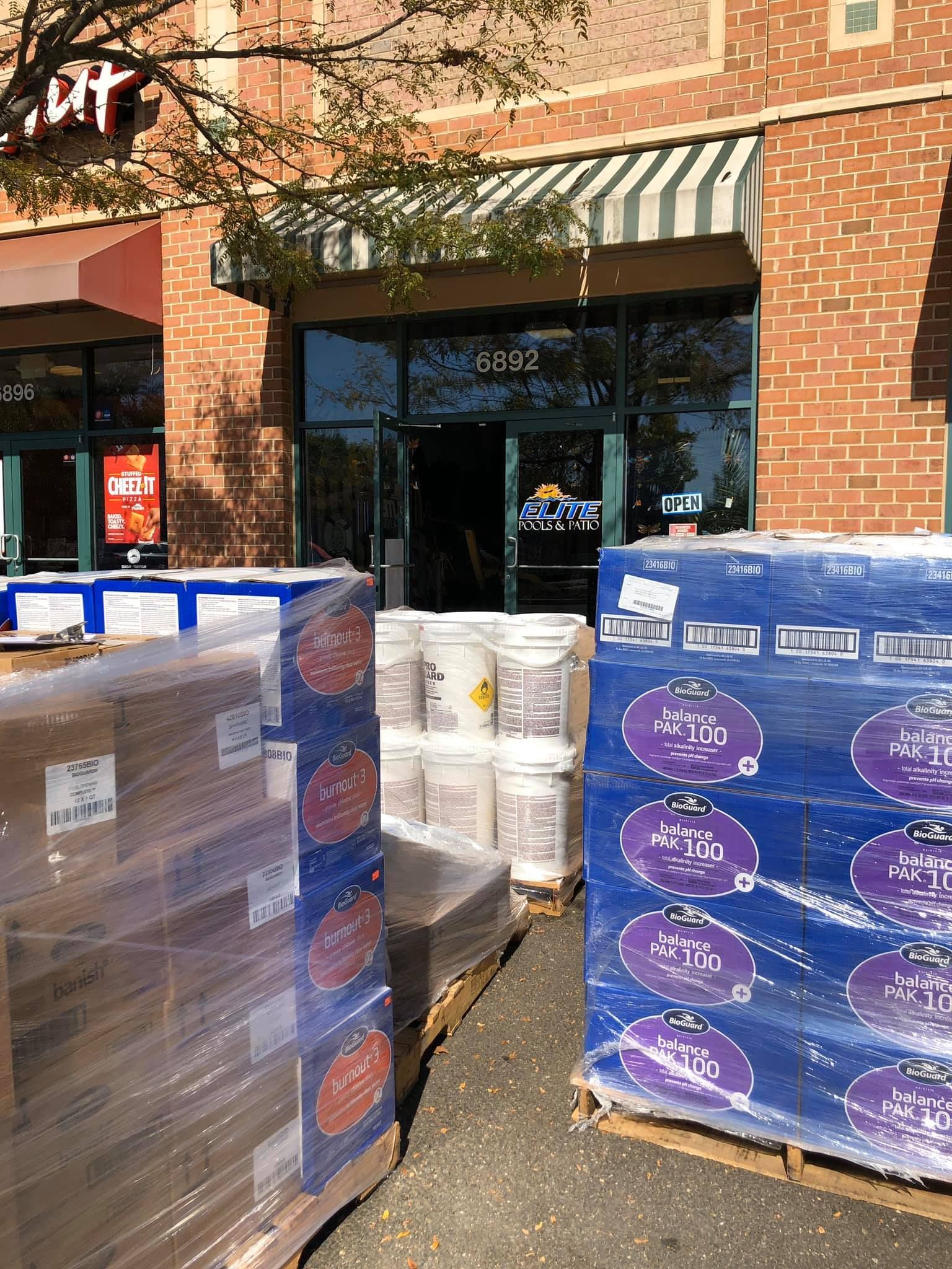 Pallets of boxes and buckets block the entrance of a store with a striped awning.