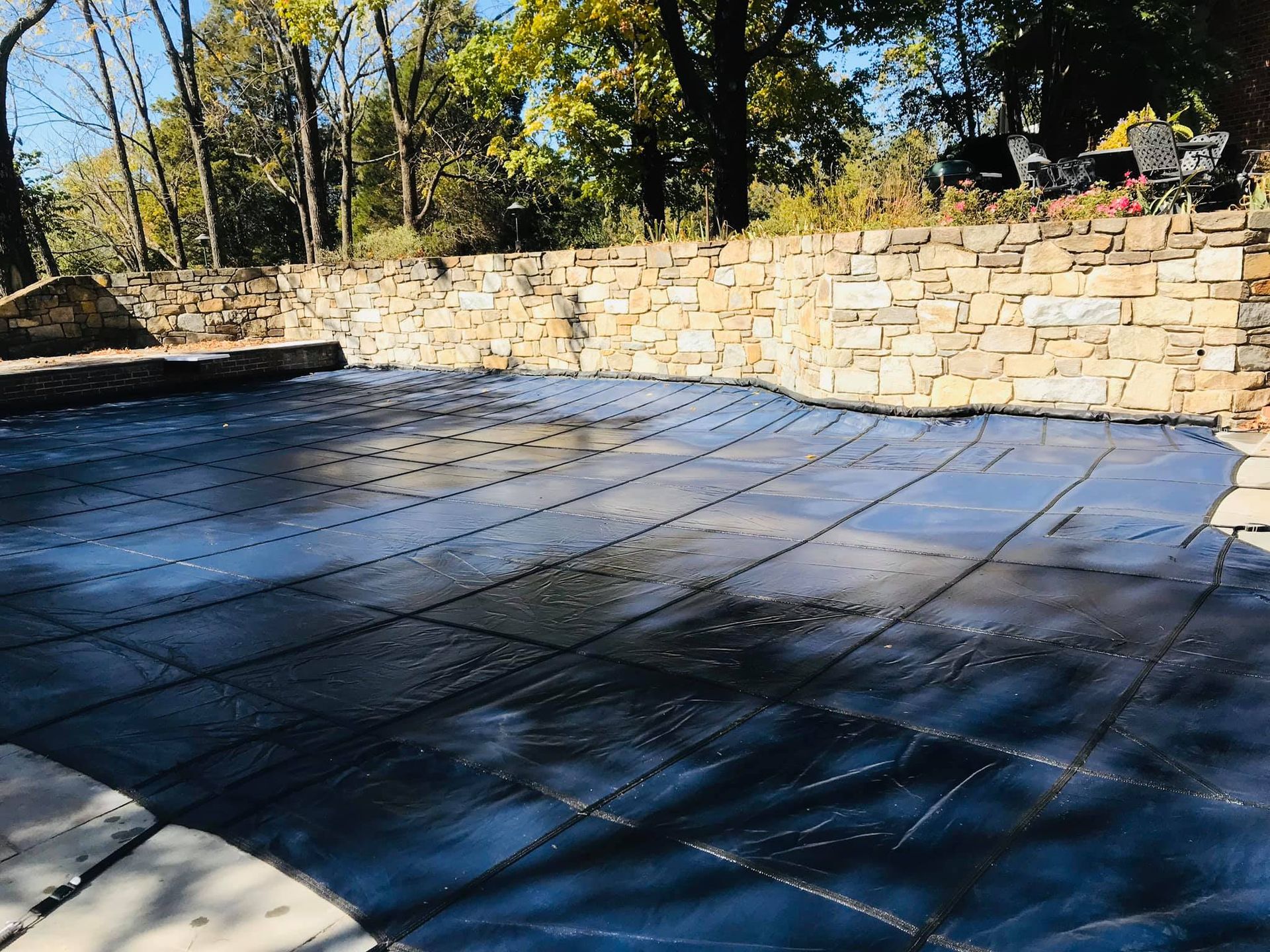 Pool covered with a dark tarp; stone wall and trees in the background.