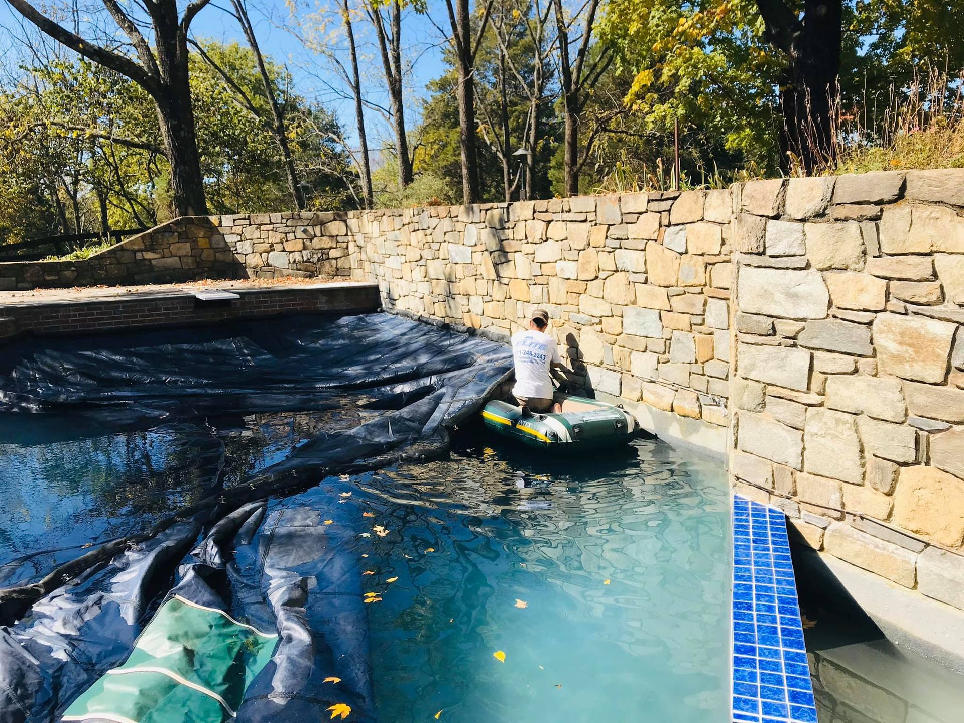 Man in pool on raft, cleaning a pool, stone wall background, trees.