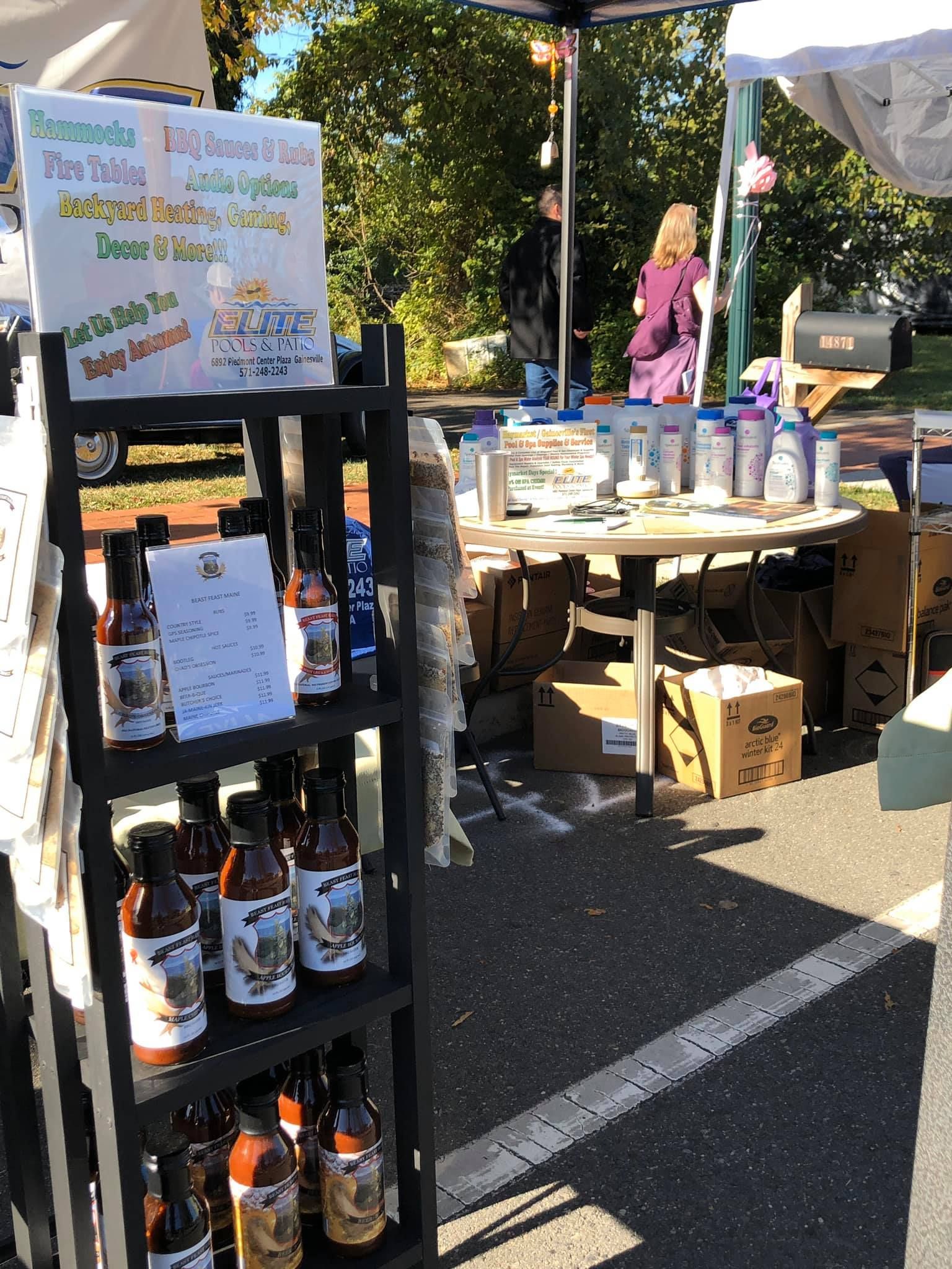 Bottles of barbecue sauce for sale at a farmer's market, with a sign and customers visible.