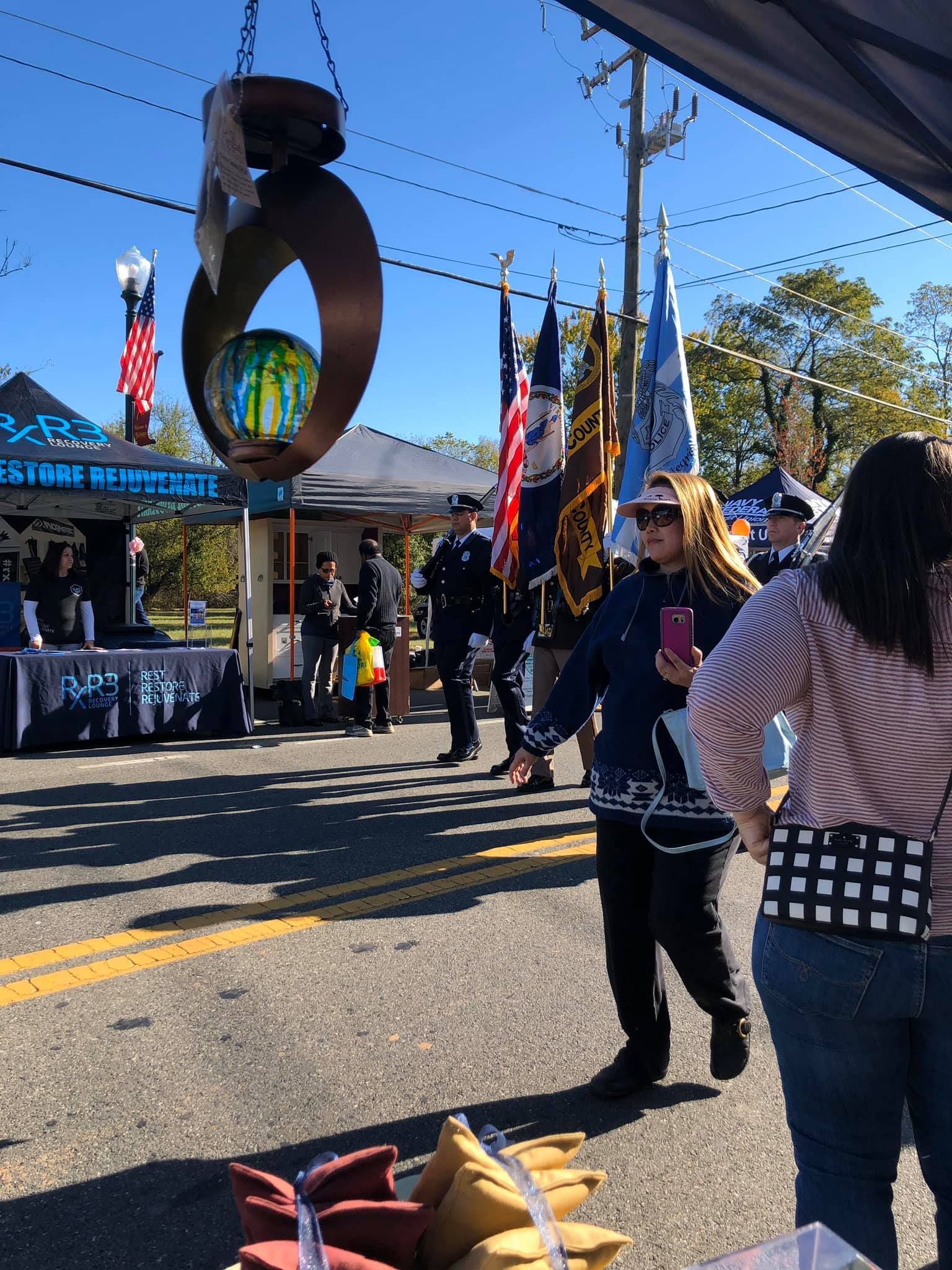 People at an outdoor event with vendor booths, flags, and an abstract metal sculpture; clear blue sky.