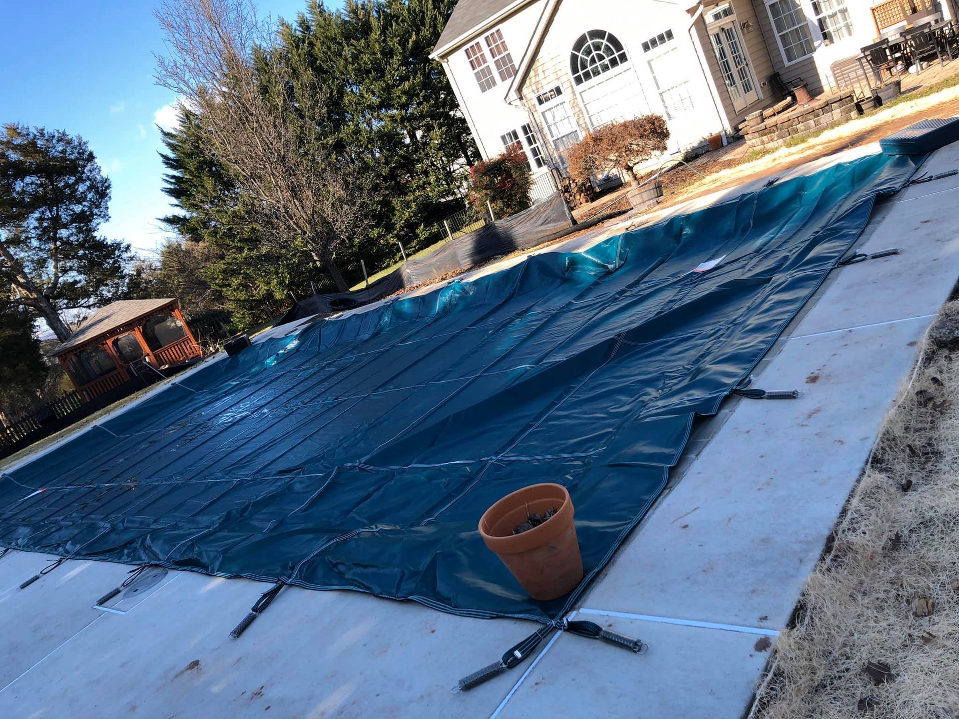 A covered pool, with a blue tarp, in front of a house. A pot sits on the concrete.