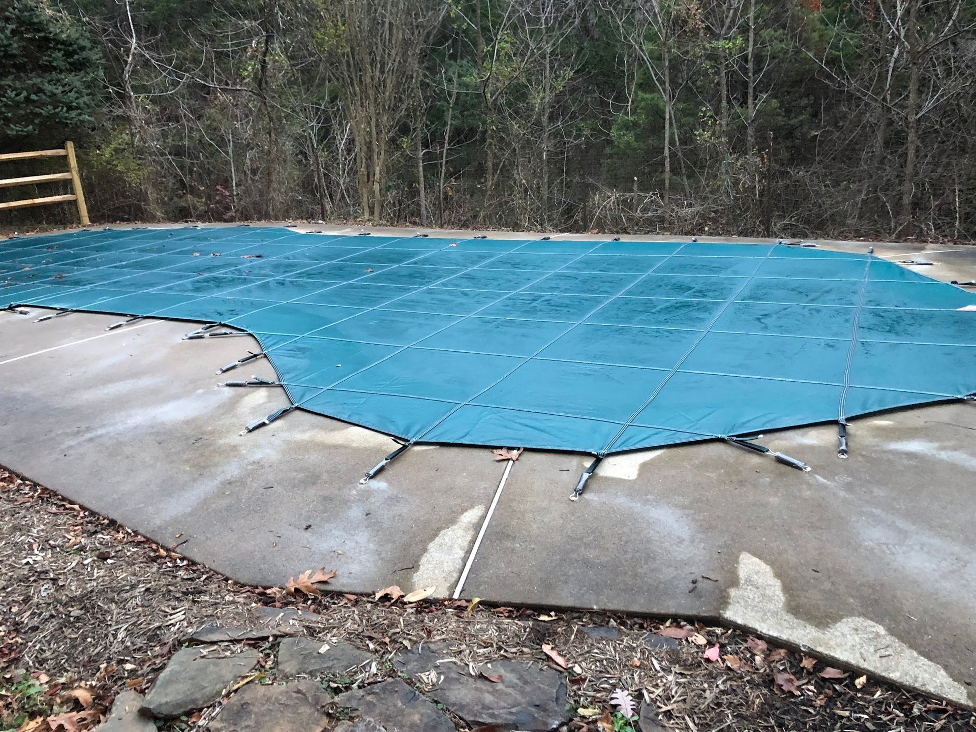 Blue pool cover stretched over a concrete patio; background of trees.