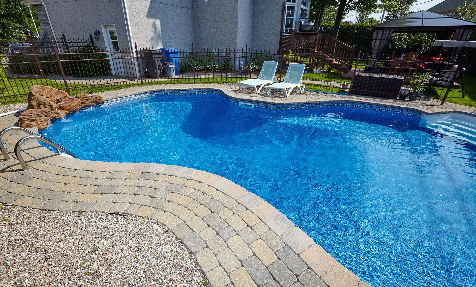 A blue-tiled swimming pool with brick edging. Two lounge chairs sit poolside, a house and gazebo in the background.
