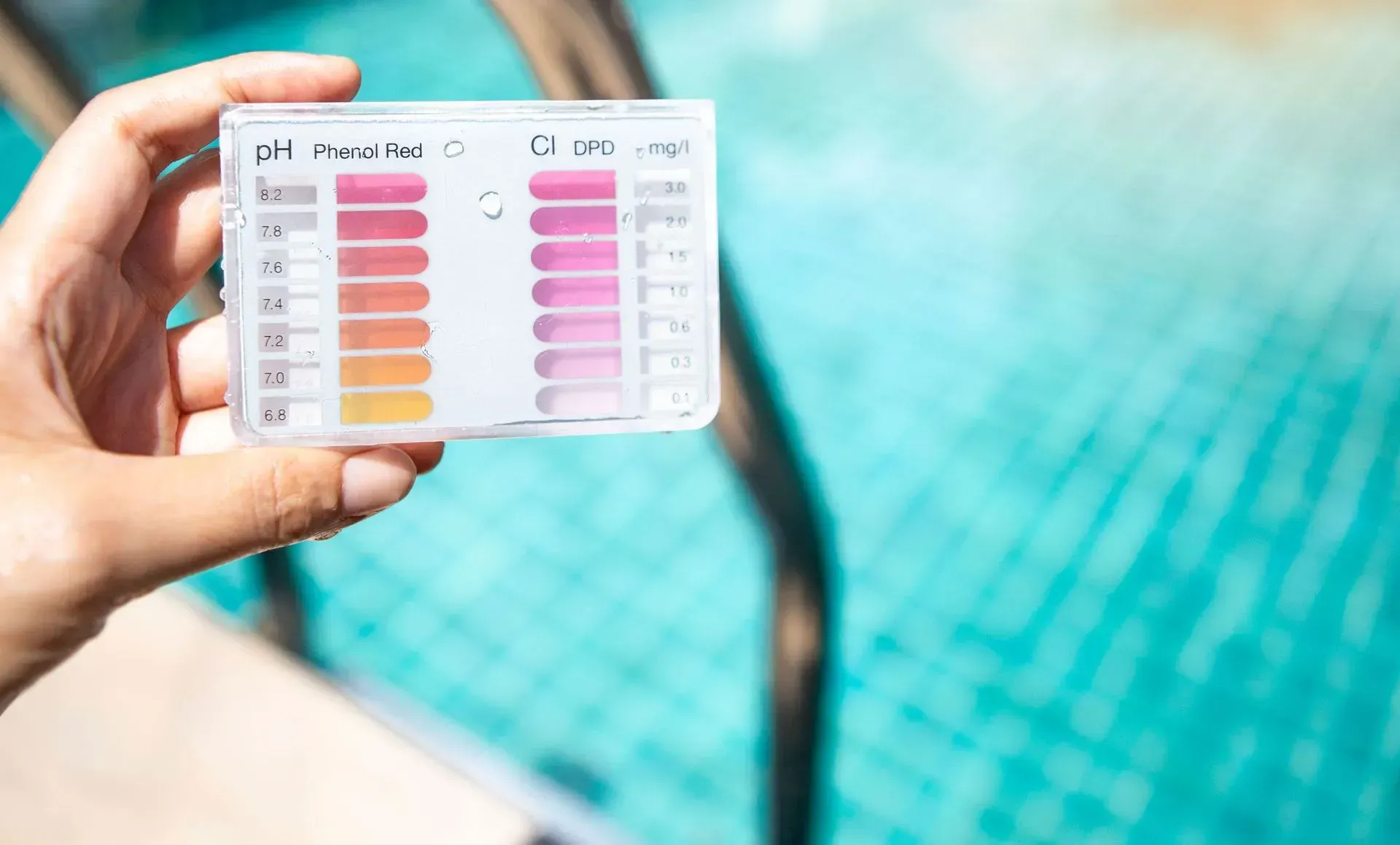 Hand holding a pool water test kit with strips showing pH and chlorine levels against a blurred pool.