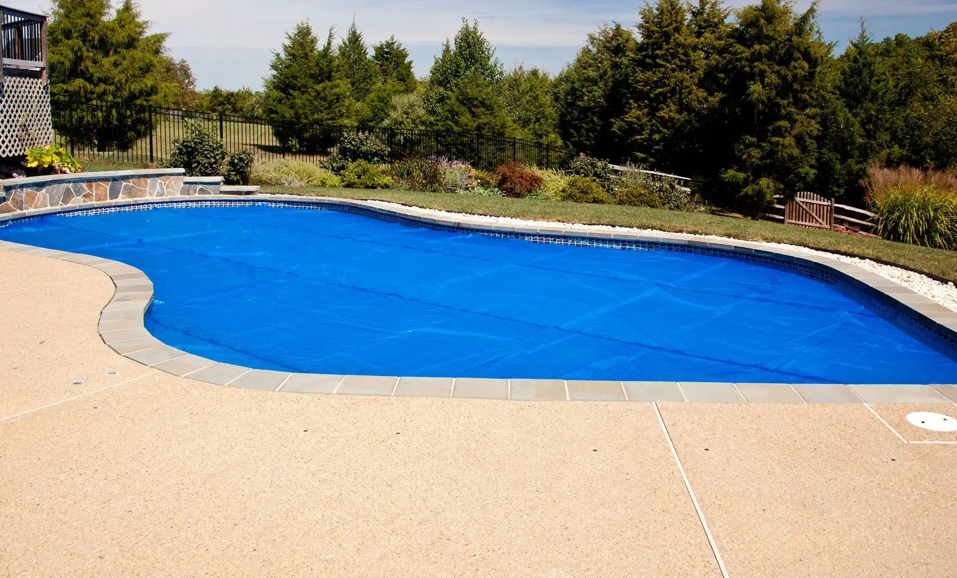 Blue pool cover over an outdoor pool with beige concrete surrounding it, trees in the background.