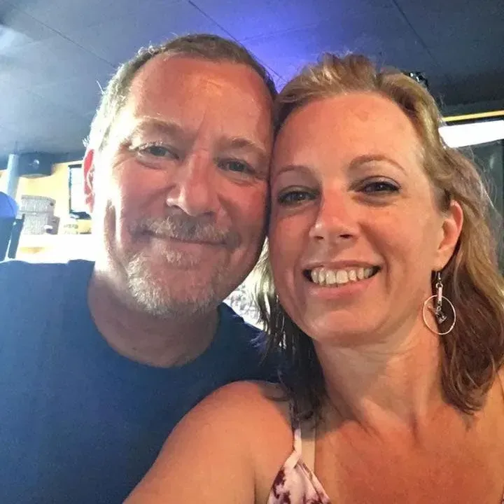 Man and woman smiling, close-up selfie. Inside, potentially a bar, with blue ceiling and lights.