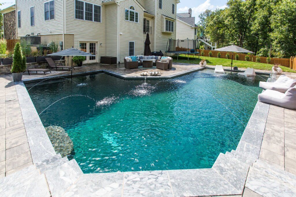 A large, dark blue pool with fountains in front of a multi-story beige house and patio furniture.
