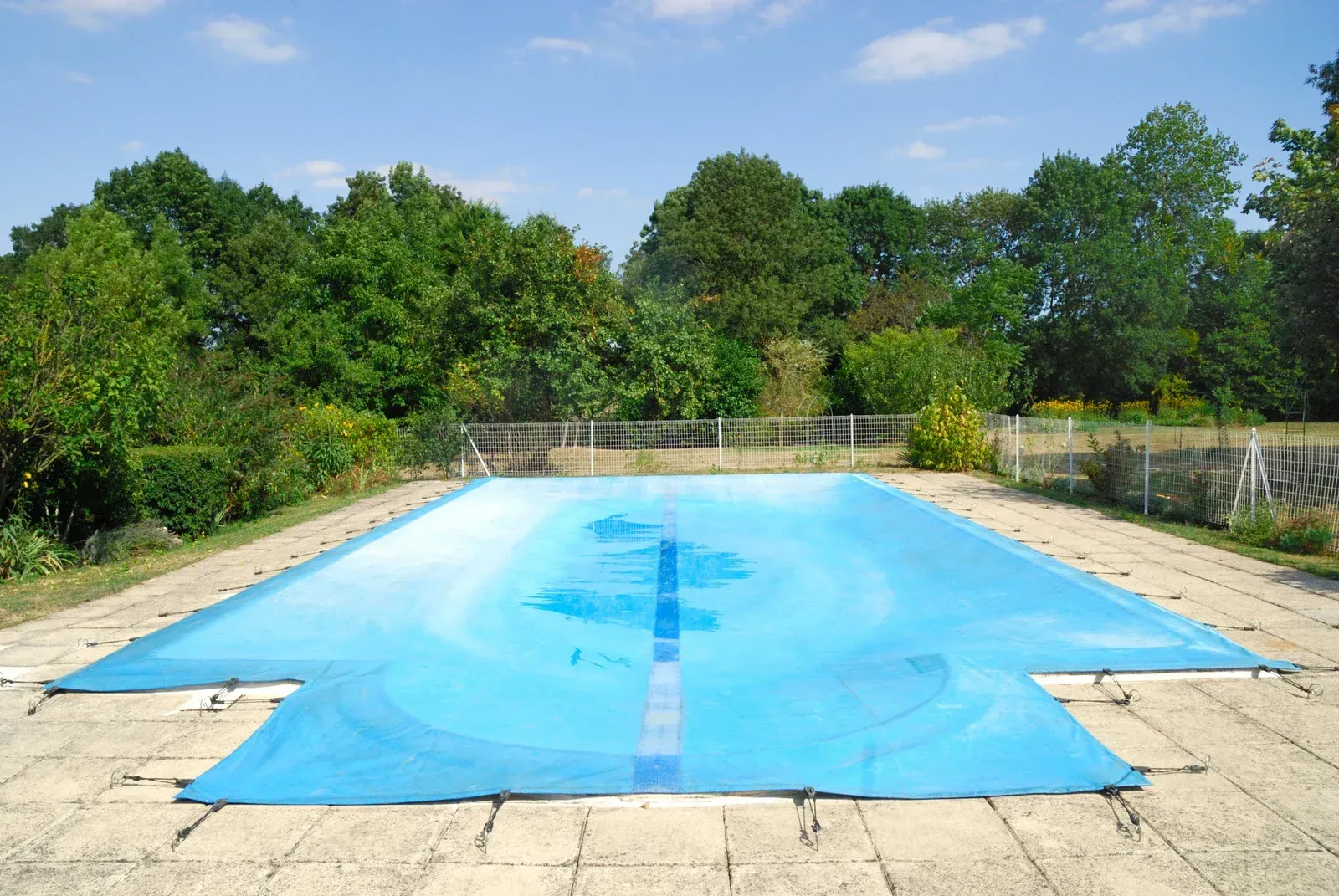 Blue pool cover over a rectangular pool, surrounded by stone paving and trees.
