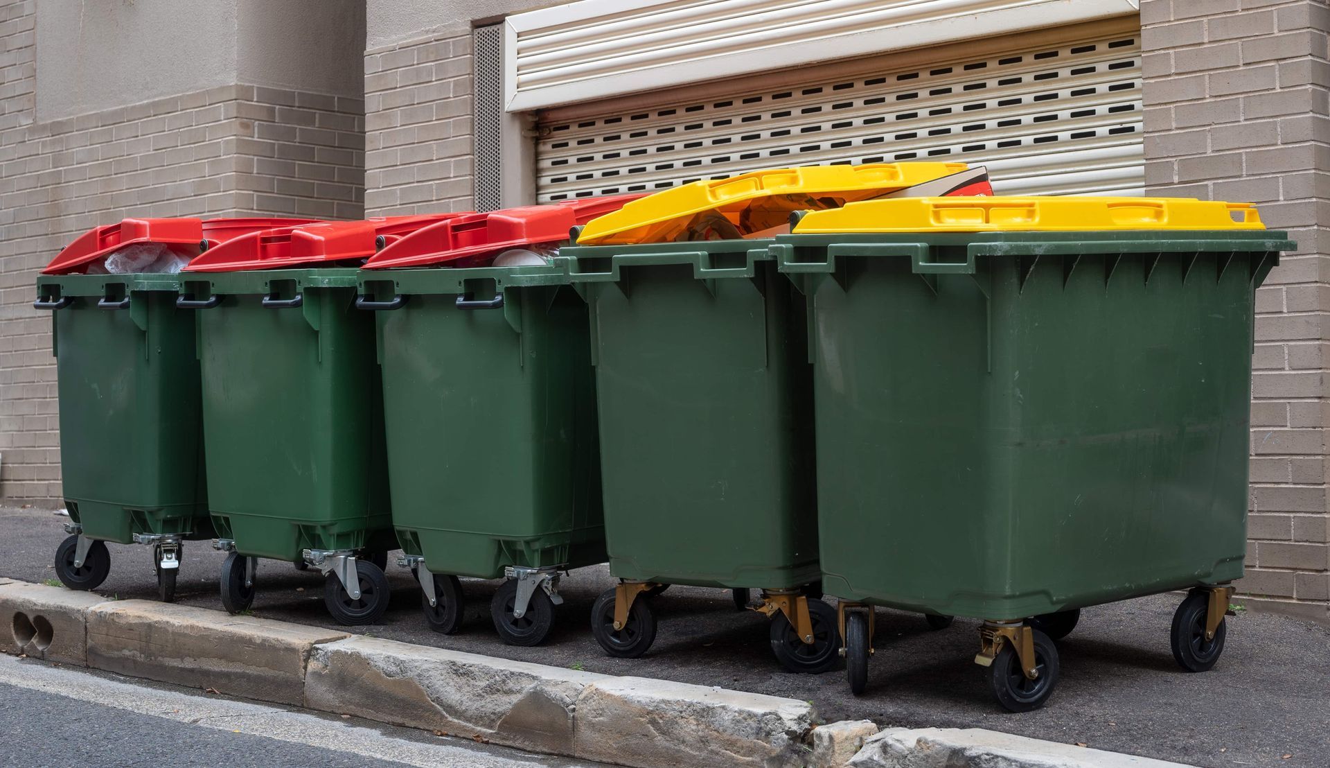 Green trash bins with red and yellow lids sit in a row next to a building.