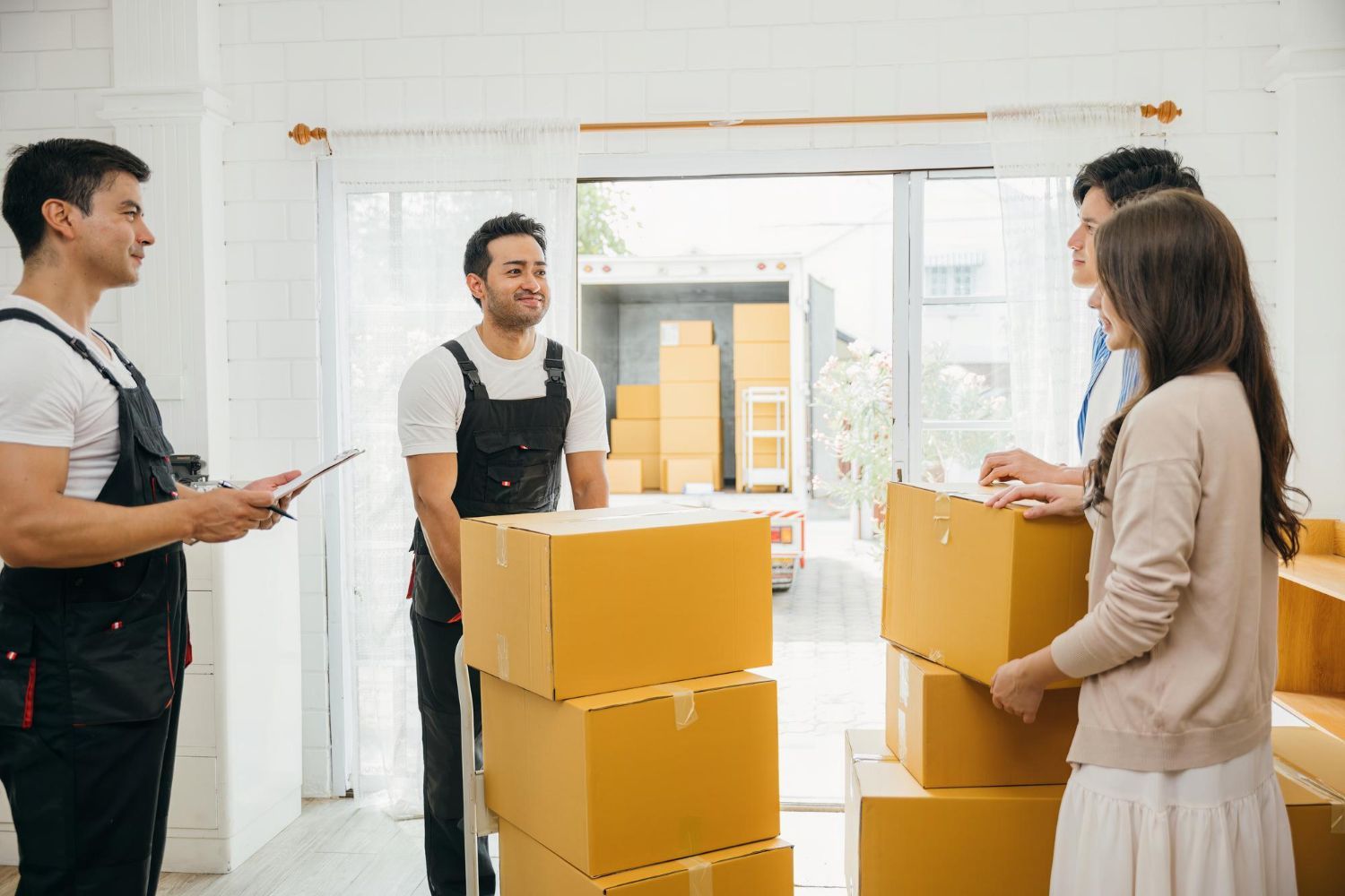Movers and a couple load boxes into a truck, inside a brightly lit room with an open doorway.
