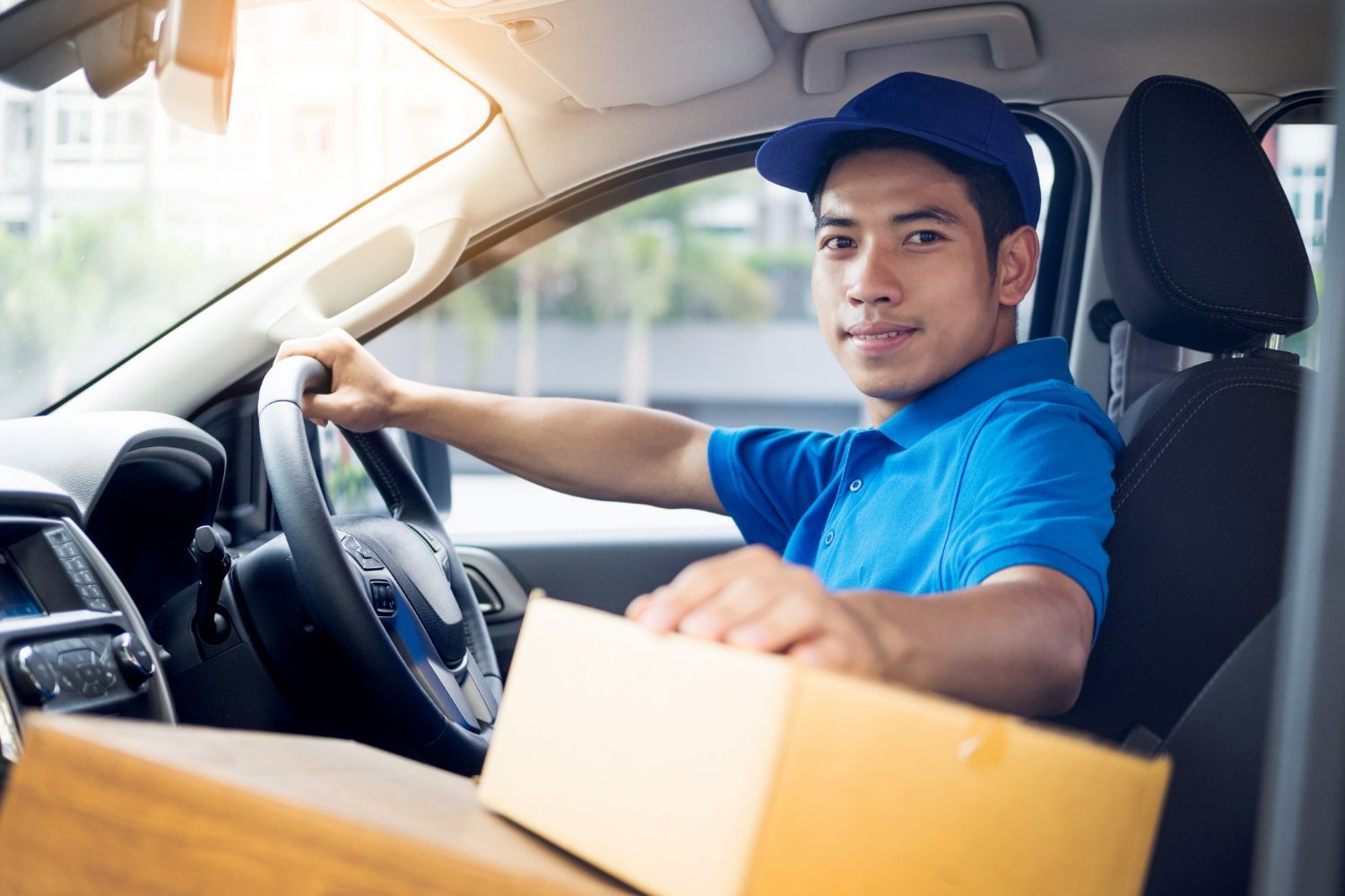Delivery driver in blue uniform and cap, smiling while holding a package in a vehicle.