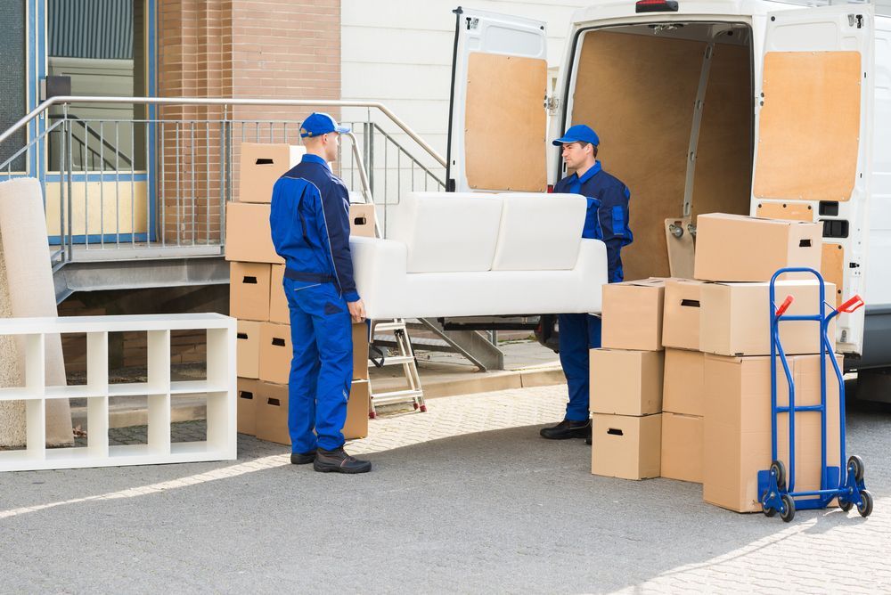 Two workers in blue uniforms move a white couch near a moving truck with stacked boxes