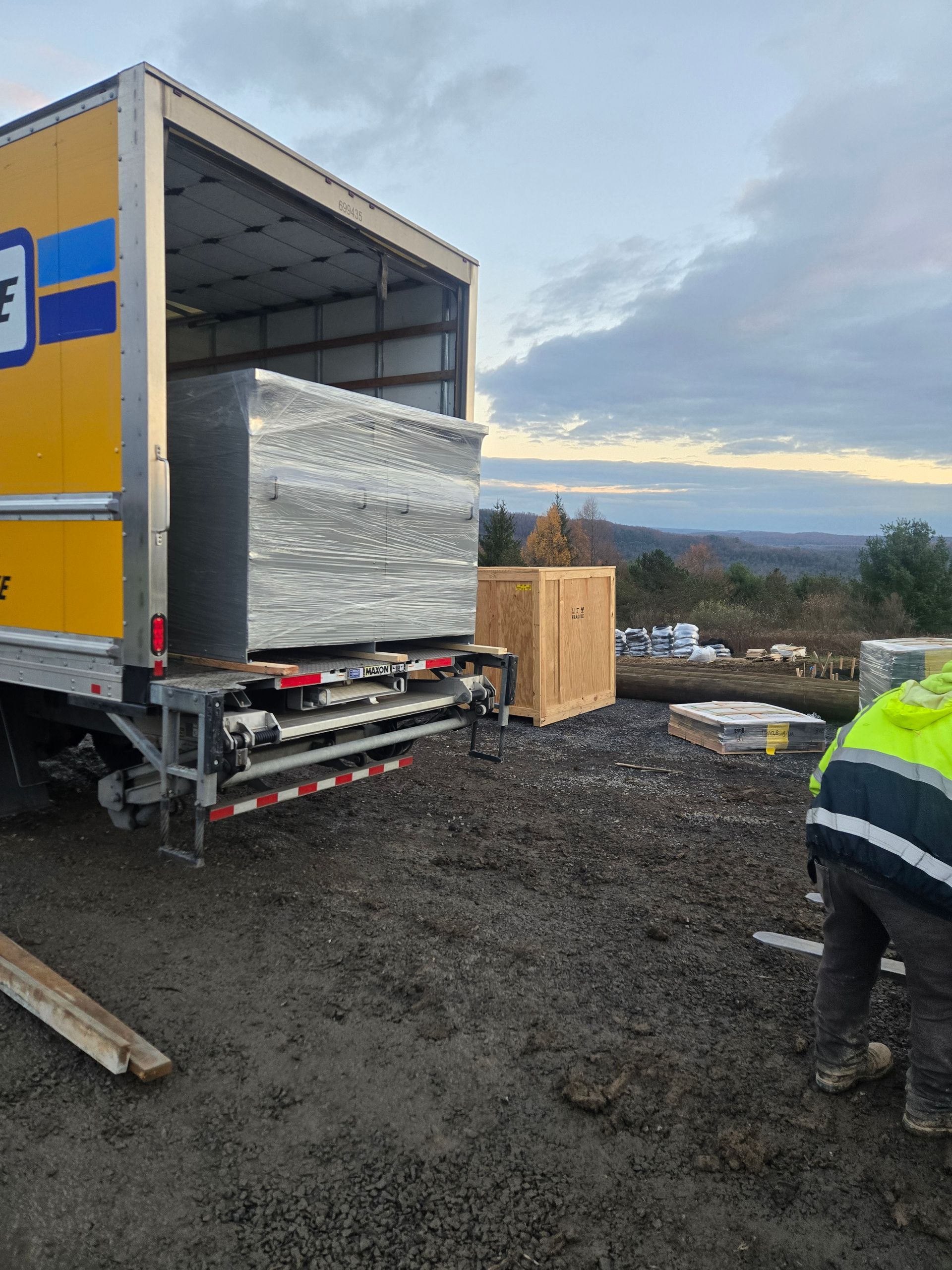 A large box being unloaded from a U-Haul truck onto a muddy ground with a worker in a high-visibility vest.