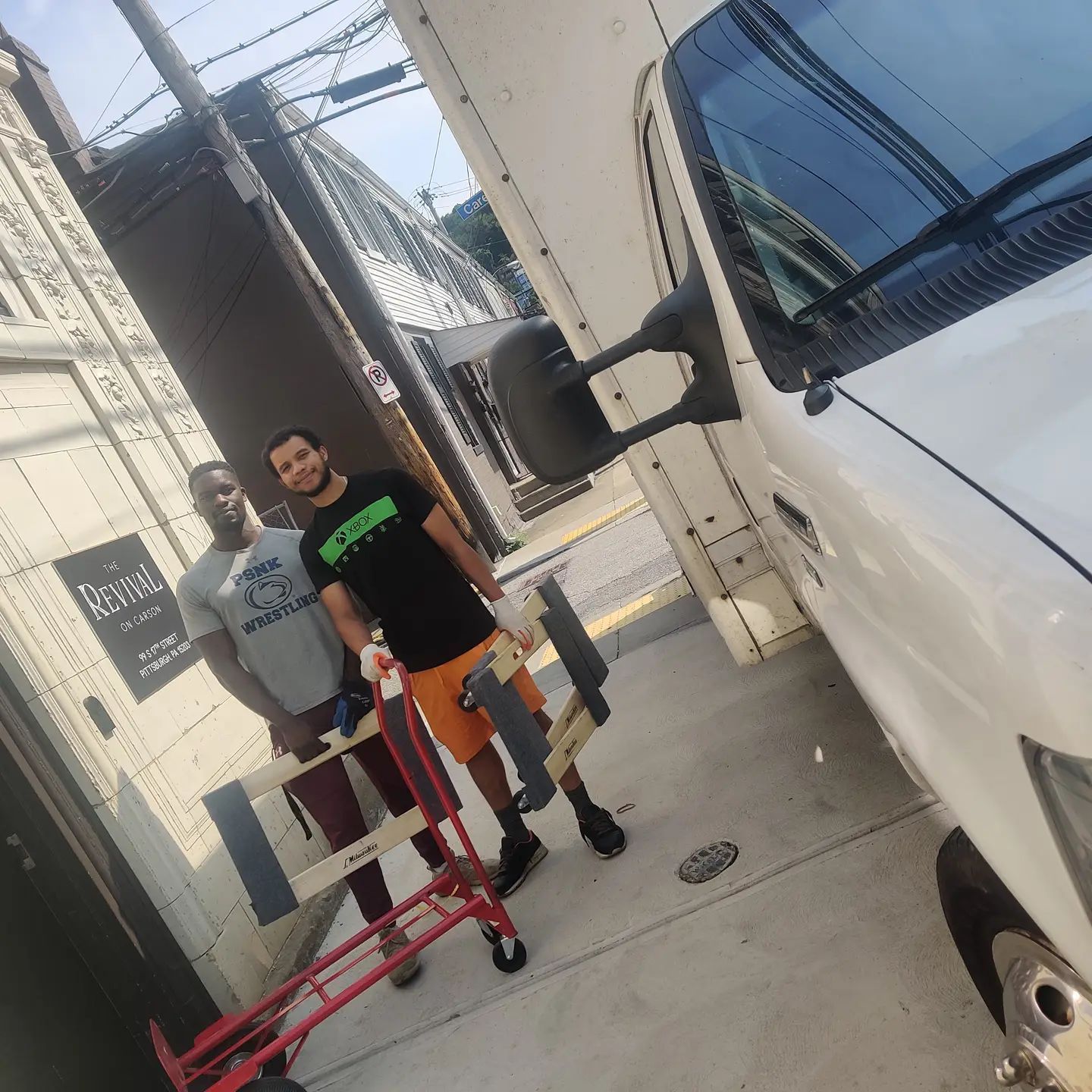 Two men standing by a white moving van. One is holding a hand truck. Sunlight.