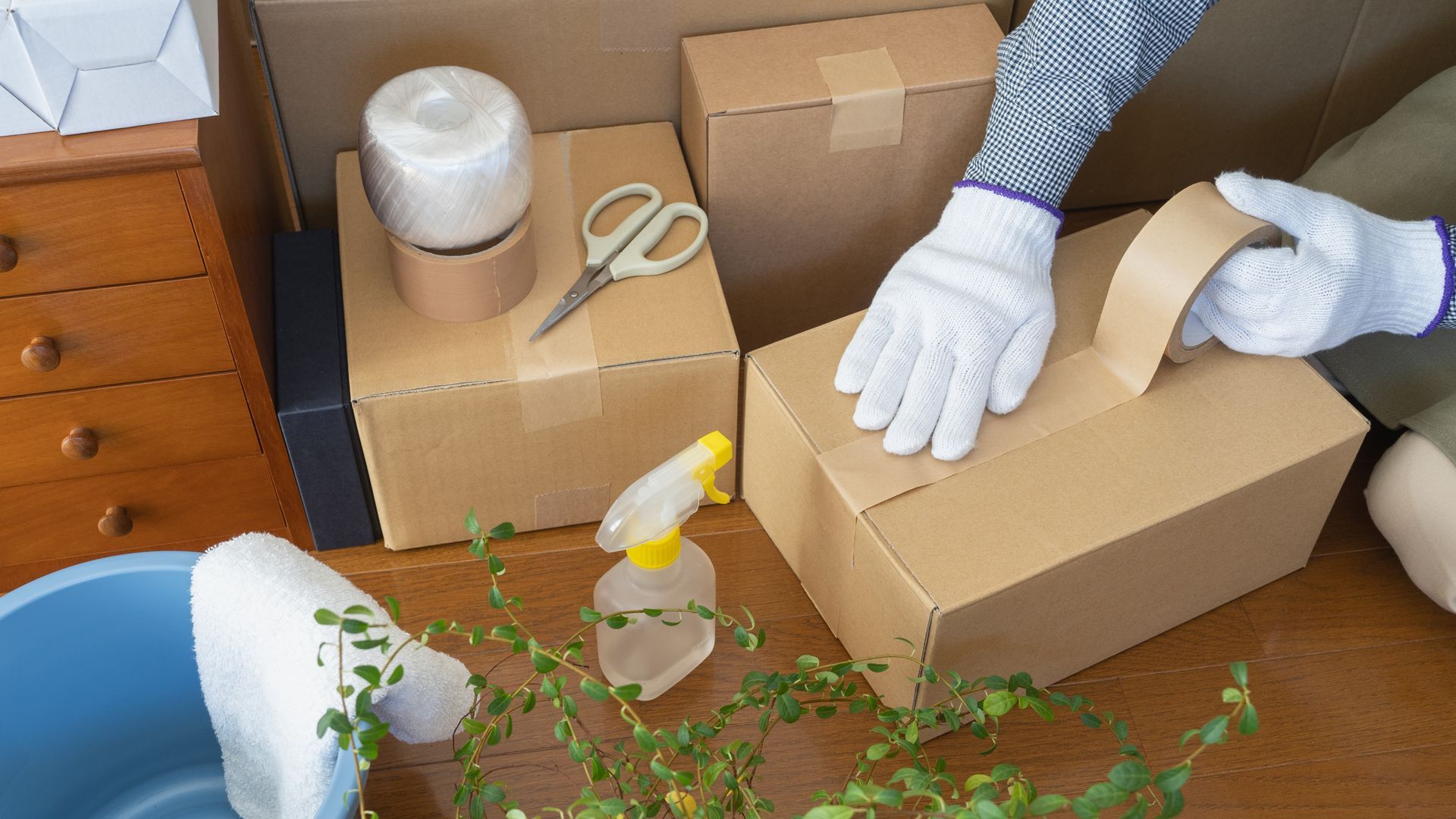 Person sealing a cardboard box with tape, with gloves on. Other boxes, scissors, tape, and a spray bottle are present.