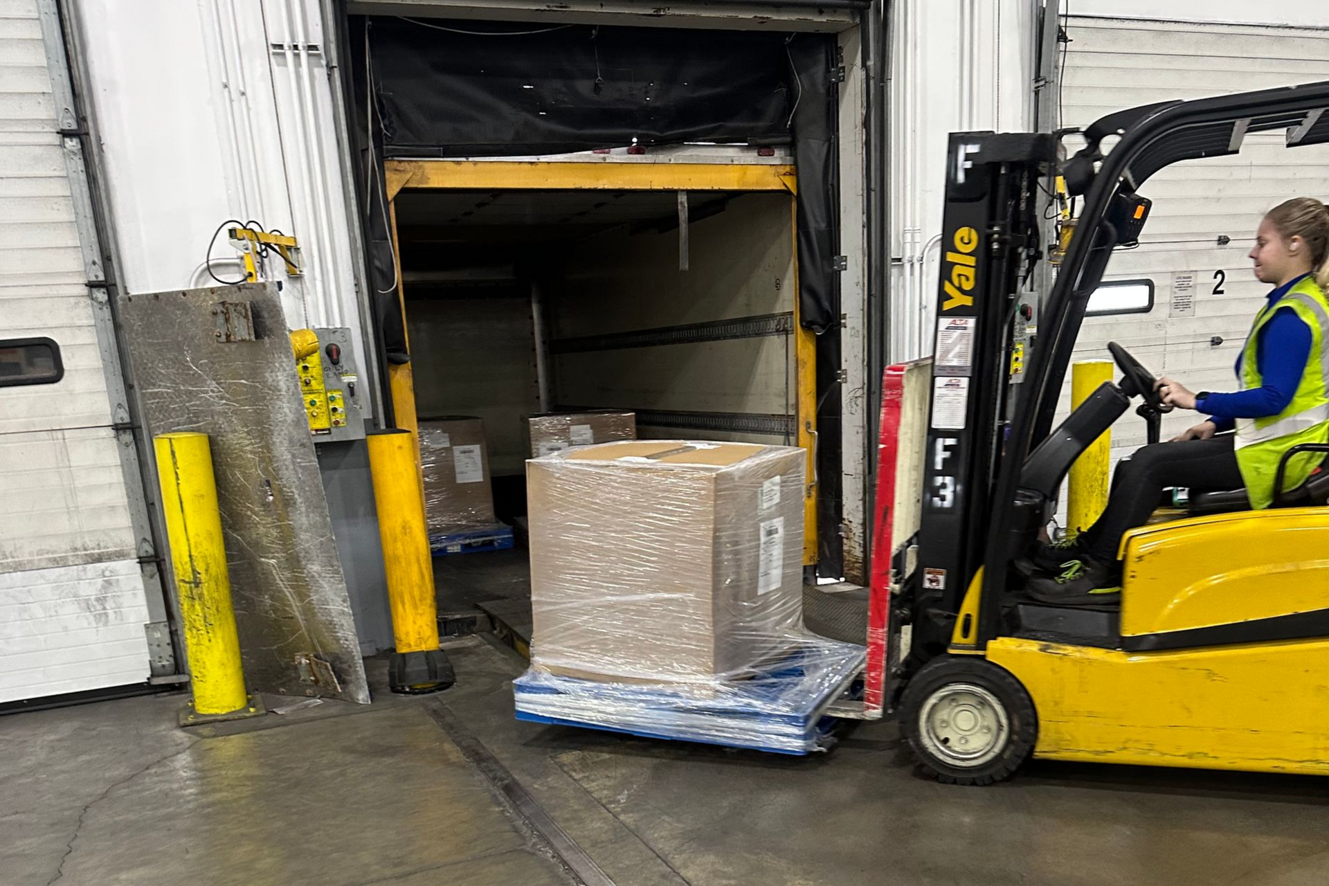 A forklift operator loads a pallet of boxes into a warehouse doorway.