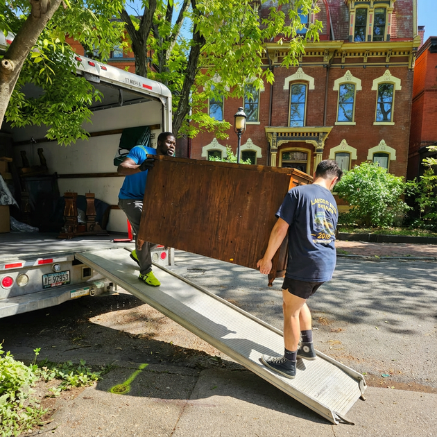 Workers unloading a large wooden panel from a truck beside a brick building