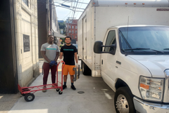 Two people stand beside a red cart on a narrow alley next to a white delivery truck.