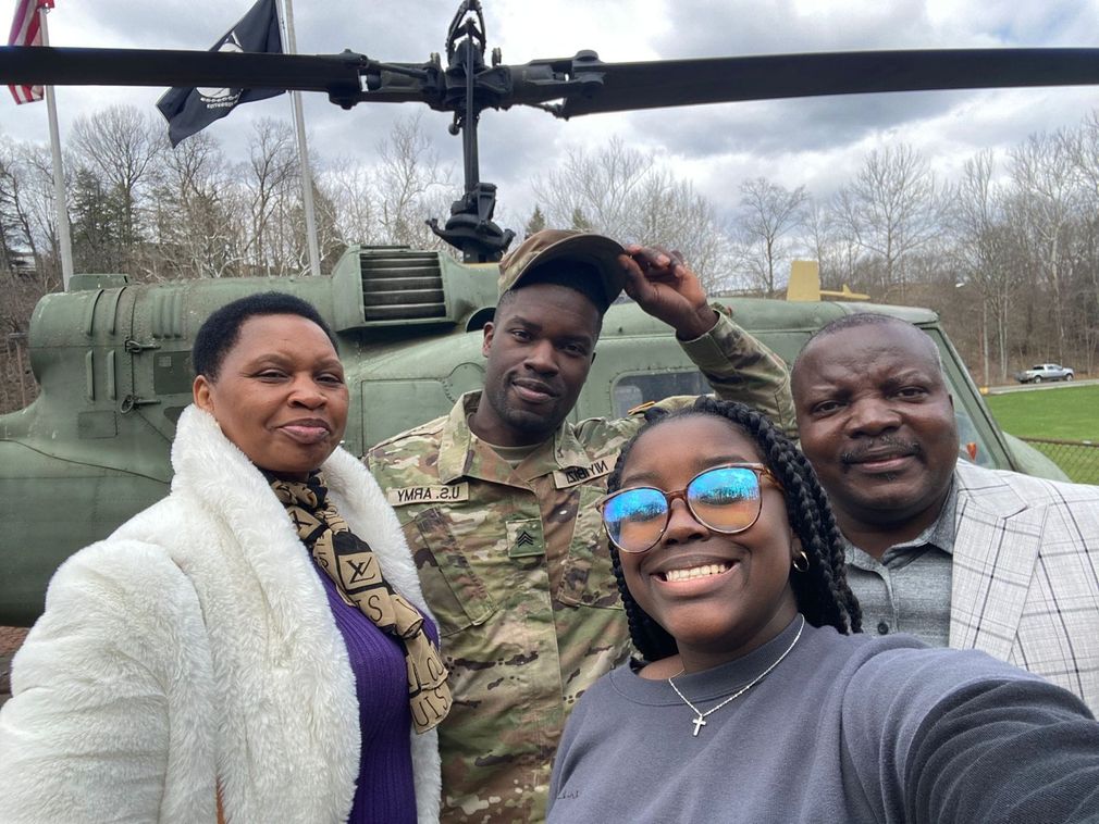 Four people pose in front of a military helicopter outdoors, one smiling and one in camouflage saluting.