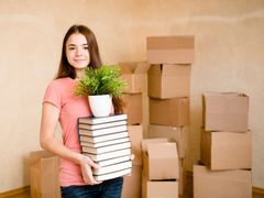 Woman holding books and a potted plant in a room with moving boxes