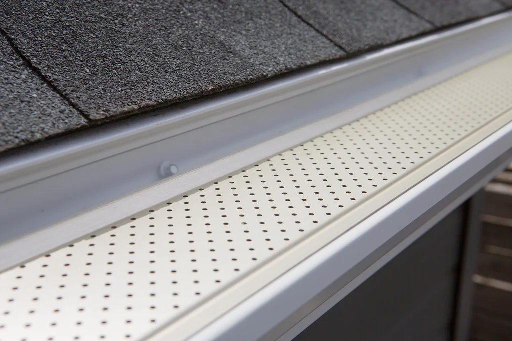 Une grille de protection de gouttière blanche perforée, installée sur une gouttière le long du bord d'un toit en bardeaux.