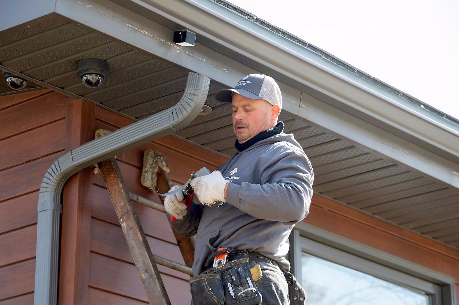 Un homme sur une échelle installe une caméra de sécurité près de la gouttière d'une maison aux murs bruns.