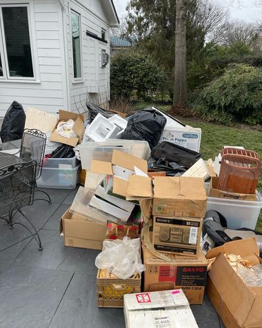 Piles of cardboard boxes and black trash bags stacked outside a white house on a patio