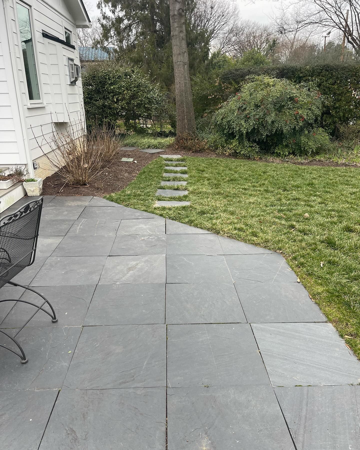 Stone patio beside a white house, leading to a grassy yard with stepping stones and shrubs.