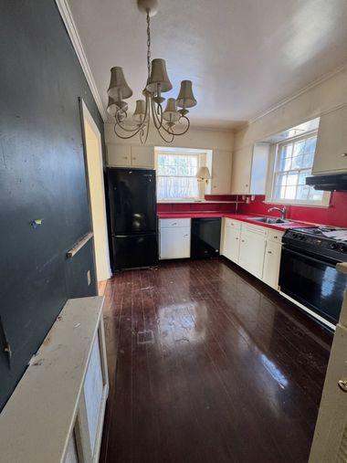 Narrow kitchen with dark wood floor, black fridge and appliances, red backsplash, and chandelier.
