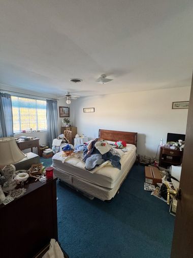 Messy bedroom with an unmade bed, blue carpet, and a window with curtains on the left.