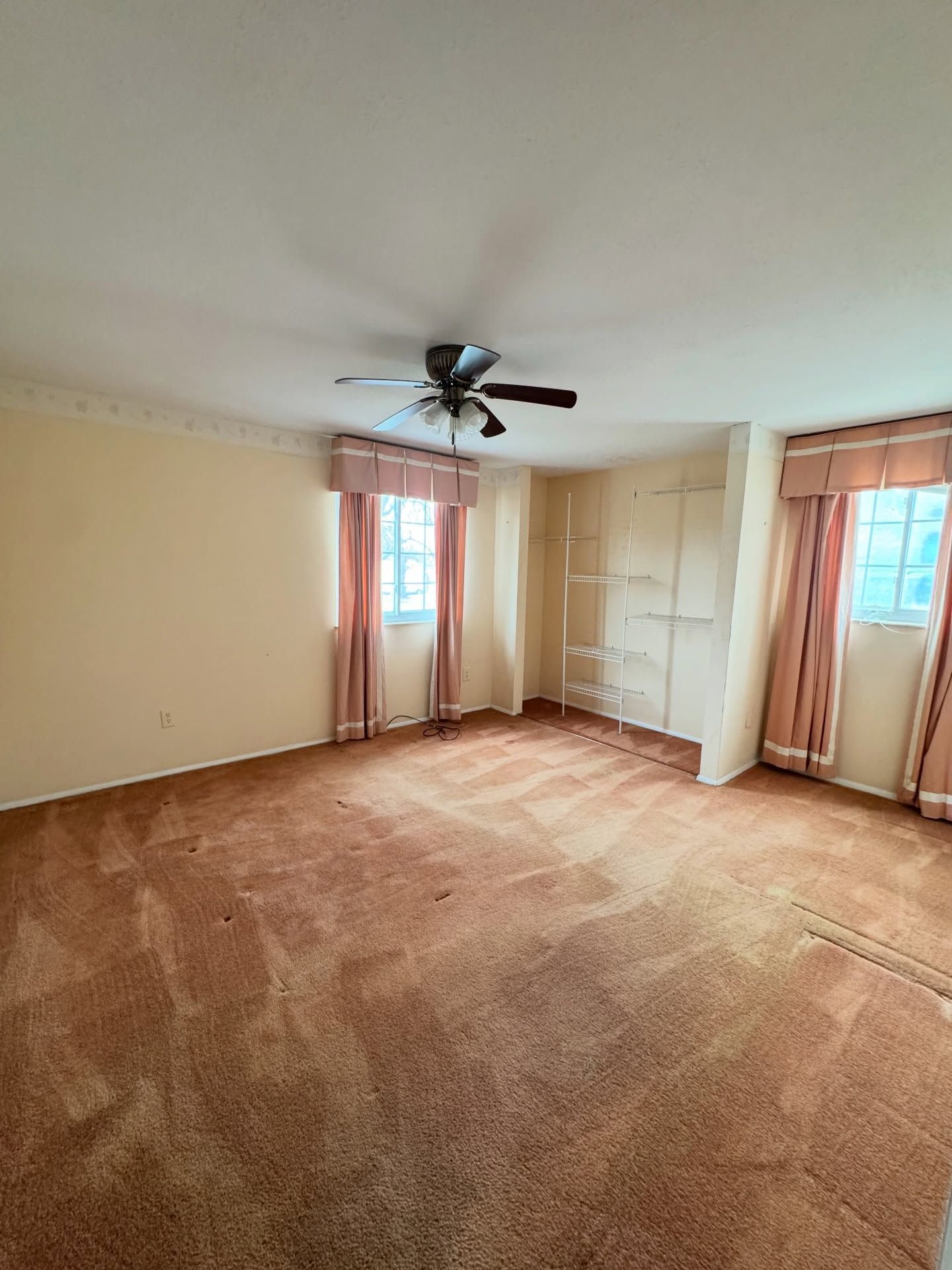 Empty beige bedroom with carpet, ceiling fan, windows with curtains, and built-in shelves