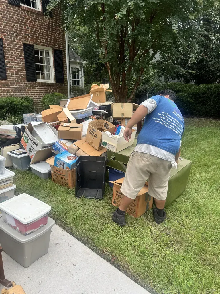 Person sorting a pile of boxes and household items on a lawn beside a brick house.