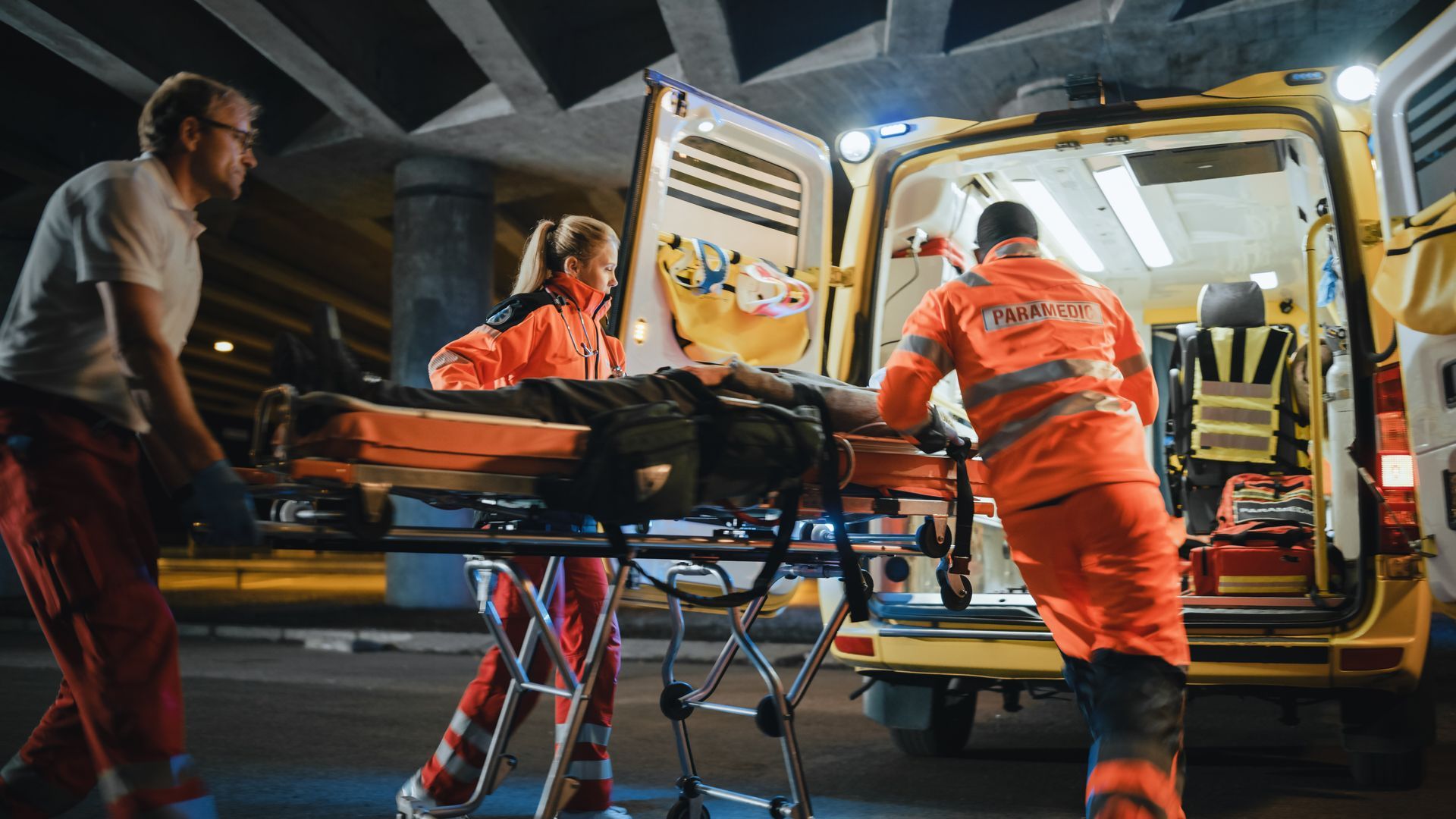 Paramedics load a patient on a stretcher into an ambulance at night, with one person assisting nearby.