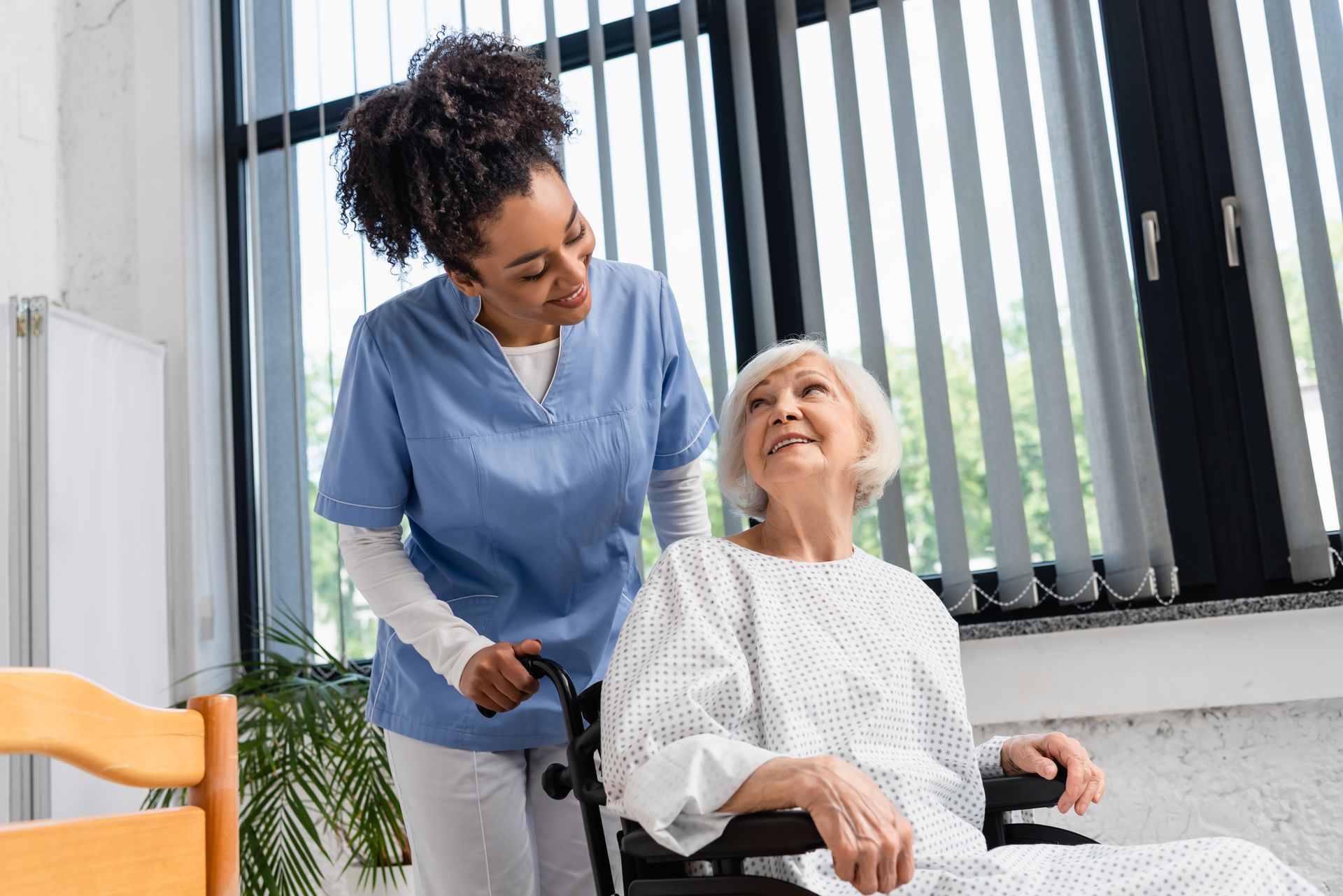 Caregiver in blue assisting a smiling elderly woman in a wheelchair indoors by a window.