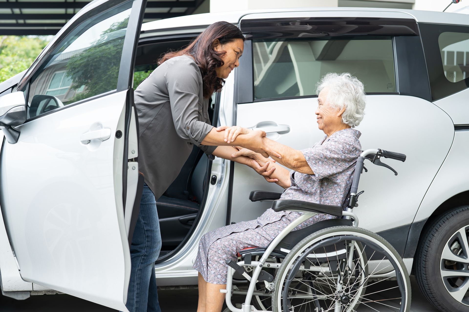 Woman helping an elderly person into a wheelchair beside a white car