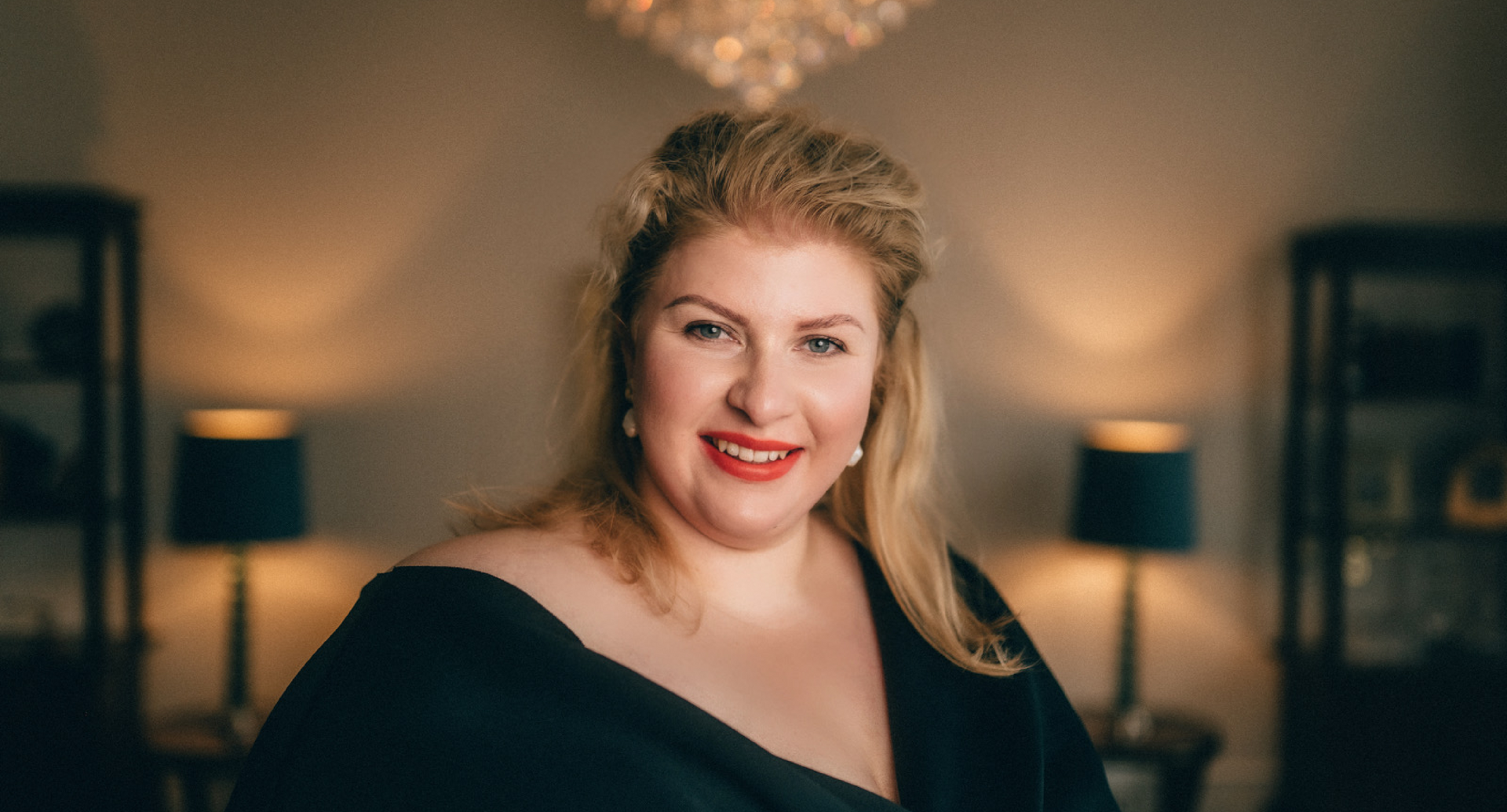 Eine Frau in einem schwarzen Kleid lächelt in einem Wohnzimmer.