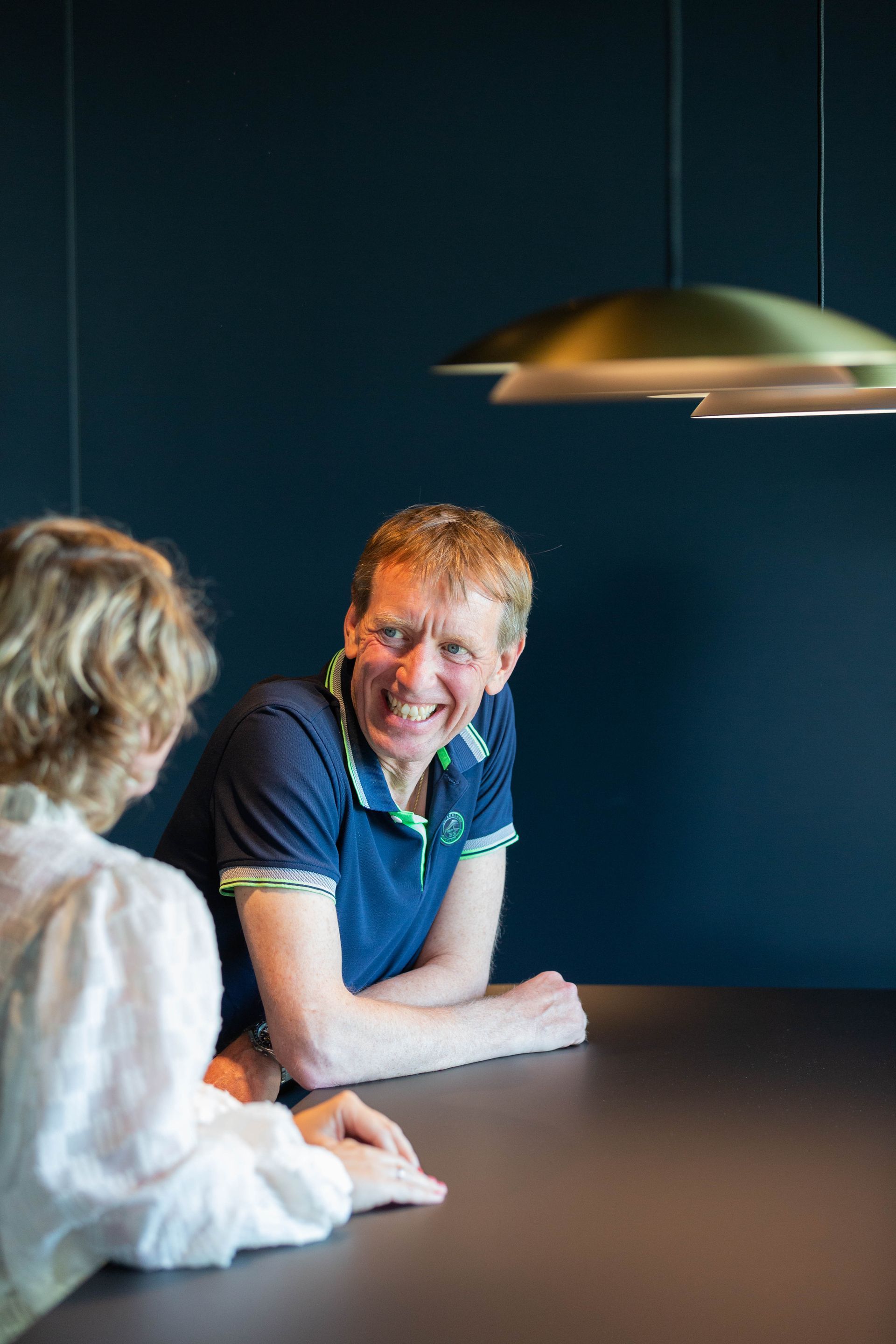 Twee mensen praten aan een tafel in een schemerig verlichte kamer onder een hanglamp.