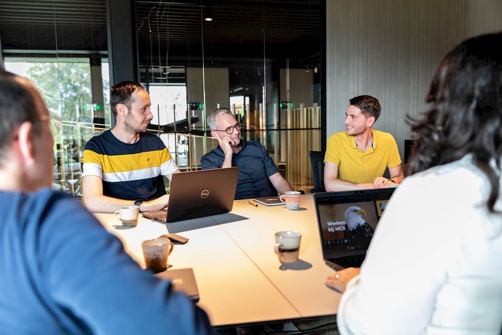 “Groep collega’s in overleg aan een vergadertafel in modern MCX kantoor.”