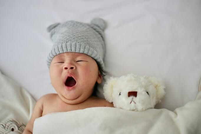 A baby wearing a hat is yawning next to a teddy bear.