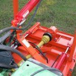 Orange tractor-mounted mower with a red hydraulic cylinder and black power shaft, on green grass.
