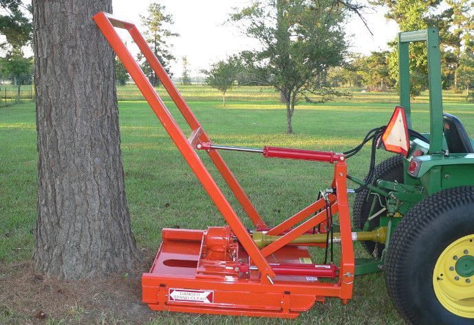 Orange tree-pulling attachment connected to a green tractor next to a tree in a grassy field.