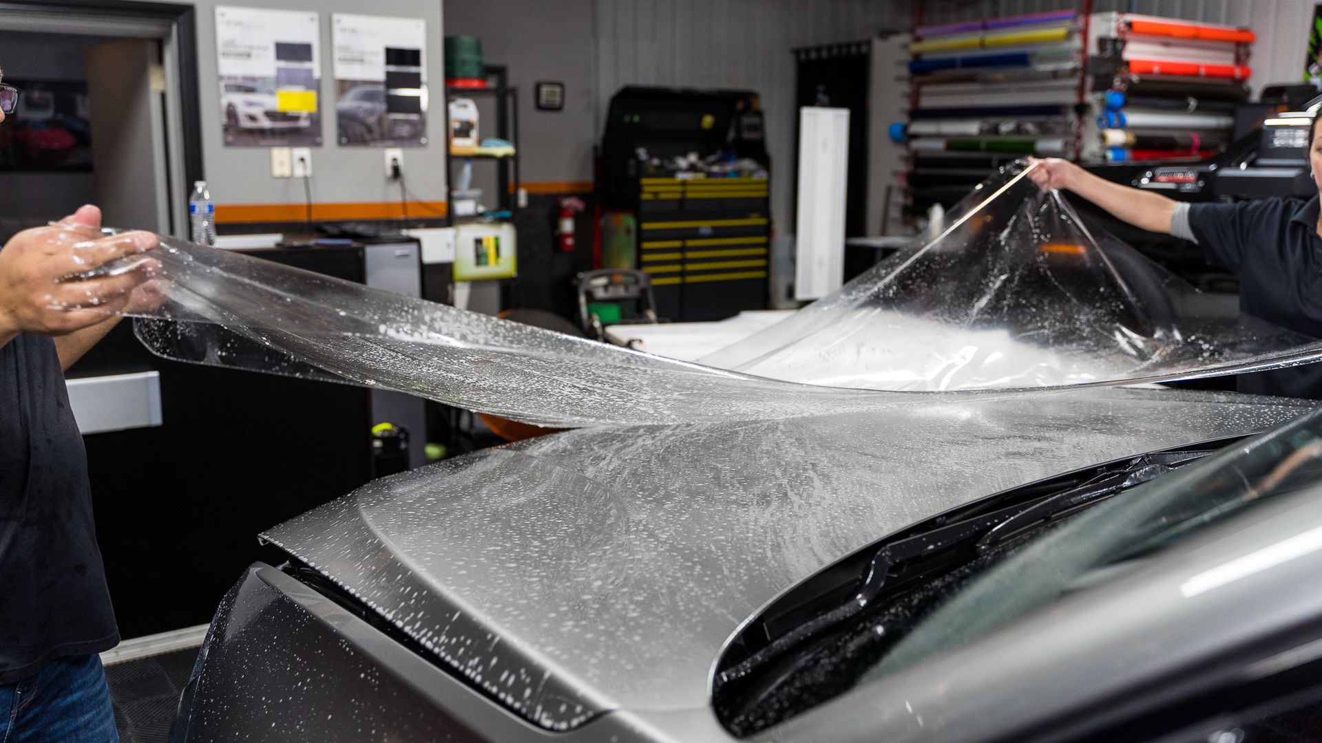 Two people applying clear protective film to the hood of a silver car inside a workshop.
